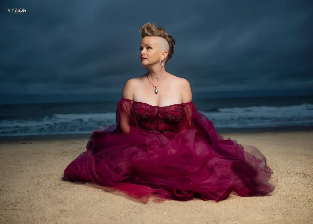 Woman in a burgundy gown seated on the beach beneath a dramatic sky